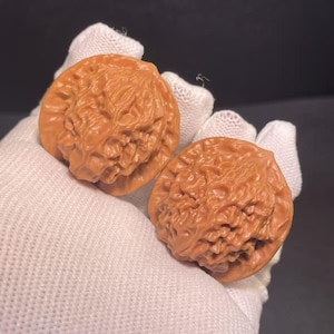 May include: Two brown, intricately carved walnuts are displayed. The walnuts have a textured surface with detailed carvings. The image is taken from a close-up perspective, highlighting the fine details of the carvings.