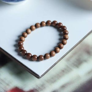May include: A brown beaded bracelet with smooth, round beads. The bracelet is made of wood and is displayed on a white surface. The beads are a warm, natural brown color. The bracelet is a simple, elegant piece of jewelry.