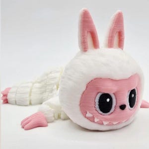 May include: A white and pink articulated creature figurine with long rabbit-like ears, large black eyes, a small nose, and visible teeth. The body features a segmented, layered design with pink hands and feet, appearing to be 3D printed.