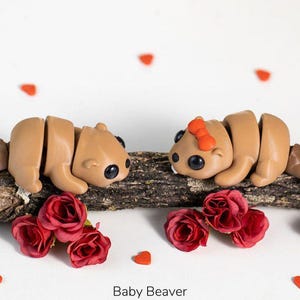May include: Two light brown articulated beaver figurines with dark eyes, one featuring a small orange bow on its head, positioned on a textured wooden branch. The scene is decorated with several small red fabric roses and tiny red heart shapes.