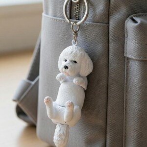 May include: A white Bichon Frise dog keychain hanging from a gray backpack zipper. The dog charm is detailed with black eyes and pink accents on its paws and tongue. The keychain is attached to a silver ring.