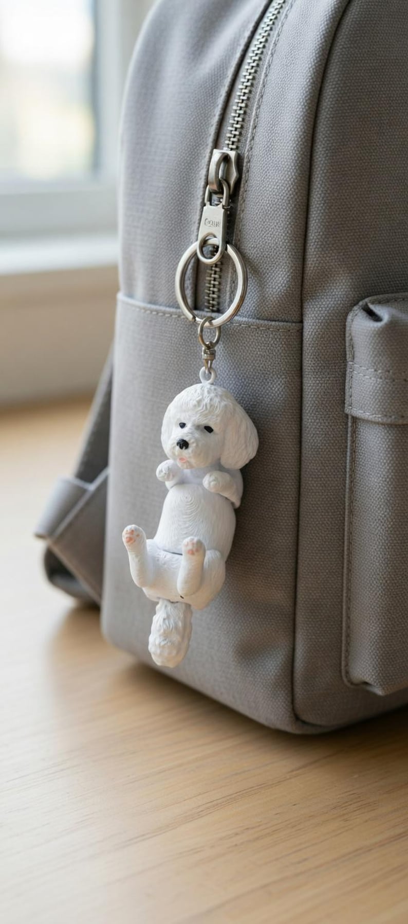 May include: A white Bichon Frise dog keychain hanging from a gray backpack zipper. The dog charm is detailed with black eyes and pink accents on its paws and tongue. The keychain is attached to a silver ring.