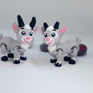 May include: Two articulated goat figurines featuring grey bodies, white chest tufts, and black horns. Each goat has large eyes, a pink nose, and black hooves with white accents. The figures are designed with movable joints at the legs and neck.