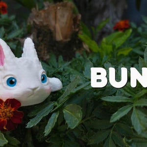 May include: A white bunny figurine with textured fur, large blue eyes, and pink inner ears, positioned among green leaves and red flowers. The text EXCLUSIVE appears in the top left corner, and the word BUNNY is printed in white on the right.