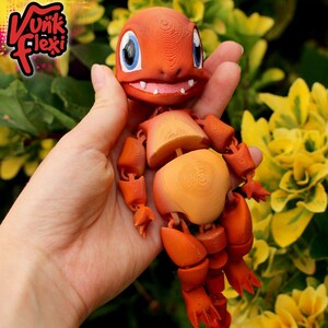 May include: An orange articulated lizard-like creature figurine with a large head, blue eyes, and white teeth. The surface shows visible 3D printed layer lines. The text Vunk Flexi is printed in the upper left corner in a stylized black font.