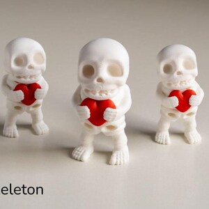 May include: Three small white skeleton figurines standing upright, each holding a bright red heart in their hands. The text "VALENTINE'S Tiny Skeleton" is printed in the lower left corner of the frame, featuring a matte finish on the figures.