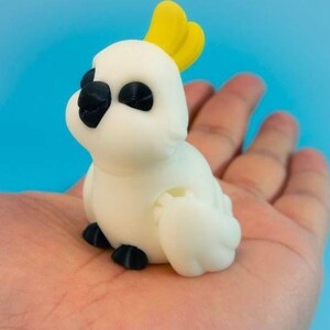May include: A small, white, 3D-printed cockatoo figurine featuring a bright yellow crest on its head, black eyes, a black beak, and black feet. The bird is depicted in a seated position with its wings tucked against its body, showcasing a smooth, matte texture throughout the surface of the object.