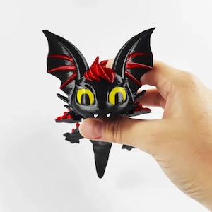 May include: A small, black, three-dimensional printed creature figurine with large, bright yellow eyes, pointed ears, and red accents on the wings and head. The figure features a smooth, glossy finish and a sharp, angular design with a pointed tail.