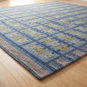 Op de afbeelding: Een blauw vloerkleed met een herhalend rasterpatroon van vierkanten in tinten olijfgroen, geel en oranje. Het kleed heeft een getextureerd oppervlak en een rand met dunne, meerkleurige horizontale strepen in rode, groene en blauwe tinten.