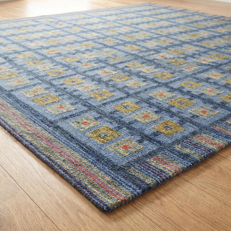 Op de afbeelding: Een blauw vloerkleed met een herhalend rasterpatroon van vierkanten in tinten olijfgroen, geel en oranje. Het kleed heeft een getextureerd oppervlak en een rand met dunne, meerkleurige horizontale strepen in rode, groene en blauwe tinten.