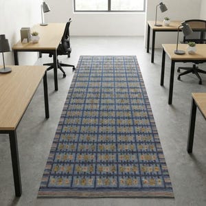 Op de afbeelding: Een lang rechthoekig vloerkleed met een blauw ruitpatroon en herhalende gouden en grijze geometrische motieven, gecentreerd op een betonnen vloer tussen rijen lichthouten bureaus met zwarte metalen frames en zwarte bureaustoelen.
