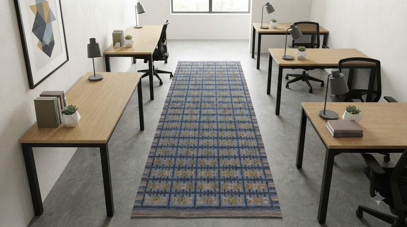Op de afbeelding: Een lang rechthoekig vloerkleed met een blauw ruitpatroon en herhalende gouden en grijze geometrische motieven, gecentreerd op een betonnen vloer tussen rijen lichthouten bureaus met zwarte metalen frames en zwarte bureaustoelen.