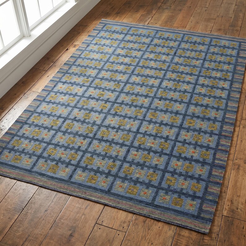 Op de afbeelding: Een rechthoekig vloerkleed met een rasterpatroon van donkerblauwe lijnen op een lichtblauwe achtergrond. Elk vierkant bevat een klein, getextureerd motief in tinten geel, groen en oranje. Het kleed heeft een gestreepte rand in blauw en beige.