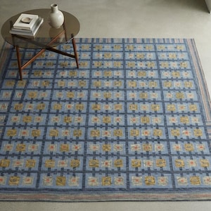 Op de afbeelding: Een groot rechthoekig vloerkleed met een rasterpatroon van donkerblauwe lijnen op een lichtblauwe achtergrond. Het raster bevat kleine vierkantjes met gele, oranje en witte bloemachtige motieven. De rand heeft dunne strepen in blauw en beige.