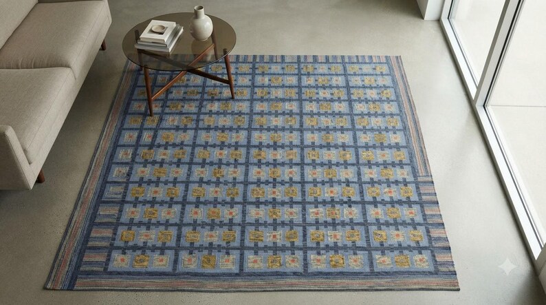 Op de afbeelding: Een groot rechthoekig vloerkleed met een rasterpatroon van donkerblauwe lijnen op een lichtblauwe achtergrond. Het raster bevat kleine vierkantjes met gele, oranje en witte bloemachtige motieven. De rand heeft dunne strepen in blauw en beige.