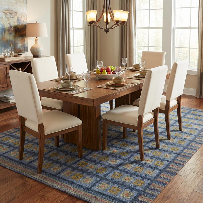 Op de afbeelding: Een rechthoekige houten eettafel met een centrale kolomvoet, omringd door zes stoelen met lichtbeige gestoffeerde zittingen en rugleuningen. De tafel is gedekt met placemats, kommen, glazen en een centrale fruitschaal.