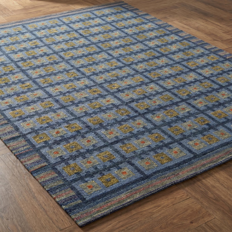 Op de afbeelding: Een rechthoekig vloerkleed met een ruitpatroon in blauwtinten. Het ontwerp bestaat uit donkerblauwe kruisende lijnen die vierkanten vormen, elk met kleine gele en oranje geometrische motieven over het gehele oppervlak.