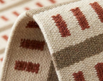 Op de afbeelding: Een geweven textiel met een crèmekleurige basis en een herhalend patroon van korte, dikke, roestkleurige rechthoekige streepjes en horizontale banden in gedempt bruin. De stof heeft een zichtbare, grove en geweven structuur.