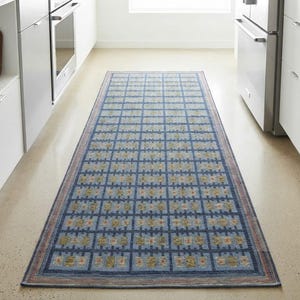 Op de afbeelding: Een rechthoekige loper met een blauwe achtergrond en een herhalend rasterpatroon van kleine, lichtgekleurde geometrische vormen en bloemachtige motieven, afgewerkt met een dunne, meerkleurige rand langs de zijkanten.