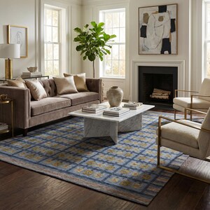 Op de afbeelding: Een groot vloerkleed met een blauw ruitpatroon en gouden en oranje bloemaccenten, geplaatst in een woonkamer met een bruine bank, twee cr&egrave;mekleurige fauteuils, een witte marmeren salontafel en een hoge groene kamerplant.