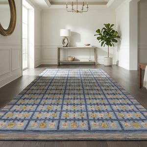 Op de afbeelding: Een rechthoekig vloerkleed met een rasterpatroon van donkerblauwe lijnen op een lichtblauwe achtergrond, met herhalende gele en oranje bloemachtige motieven in elk vierkant, afgewerkt met horizontaal gestreepte randen aan de uiteinden.