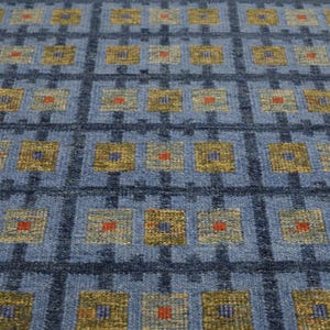 Op de afbeelding: Een geweven textielkleed met een rasterpatroon van donkerblauwe lijnen op een lichtblauwe achtergrond. Het raster bevat herhalende olijfgroene vierkanten, elk in het midden voorzien van een klein, fel oranje of blauw accent.
