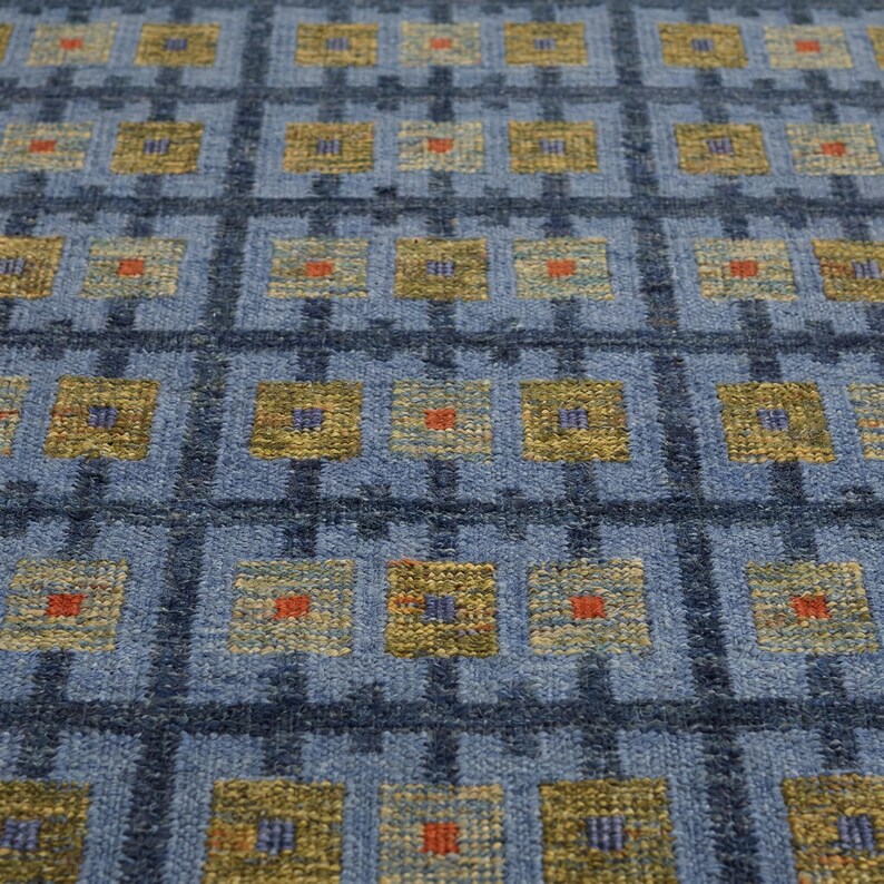 Op de afbeelding: Een geweven textielkleed met een rasterpatroon van donkerblauwe lijnen op een lichtblauwe achtergrond. Het raster bevat herhalende olijfgroene vierkanten, elk in het midden voorzien van een klein, fel oranje of blauw accent.