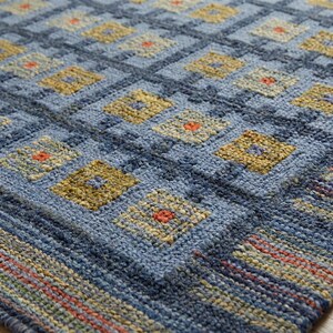 Op de afbeelding: Een getextureerd vloerkleed met een herhalend rasterpatroon van blauwe vierkanten. Elk vierkant bevat een kleine gele ruit met een centrale oranje stip. De rand toont horizontale strepen in tinten blauw, geel en oranje.