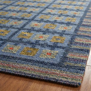 Op de afbeelding: Een geweven vloerkleed met een blauw ruitpatroon en herhalende vierkanten in olijfgroene, beige tinten en kleine oranje accenten. De rand heeft een veelkleurig gestreept ontwerp met dunne rode, groene en blauwe lijnen.
