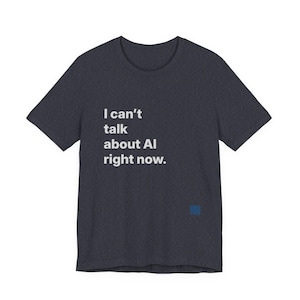 Peut inclure: T-shirt gris foncé avec le texte blanc "I can't talk about AI right now." Un petit carré bleu est dans le coin inférieur droit. Le t-shirt a des manches courtes et un col rond.
