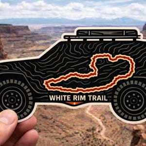May include: Black sticker featuring a stylized off-road vehicle with a roof rack and spare tire. The vehicle has a topographic design and a red outline of a trail. The text "WHITE RIM TRAIL" is displayed below the vehicle.
