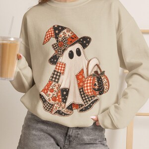 May include: A beige crewneck sweatshirt featuring a graphic of a ghost wearing a patchwork witch hat and holding a jack-o'-lantern. The ghost design incorporates various patterns including plaid, stars, and small pumpkins in orange, black, and white.