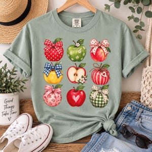Puede incluir: Una camiseta de manga corta color verde salvia con un estampado gráfico de nueve manzanas dispuestas en cuadrícula. Las manzanas están decoradas con varios patrones, incluyendo lunares, rayas, cuadros vichy y flores, cada una con un lazo decorativo.