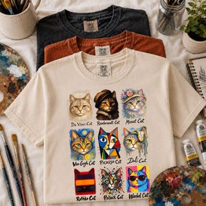 Op de afbeelding: Een crèmekleurig t-shirt met een raster van negen kattenillustraties geïnspireerd door beroemde kunstenaars, gelabeld Da Vinci Cat, Rembrandt Cat, Monet Cat, Van Gogh Cat, Picasso Cat, Dali Cat, Rothko Cat, Pollock Cat en Warhol Cat.