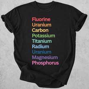 May include: A black short-sleeved t-shirt featuring a vertical list of chemical elements printed in a rainbow color gradient. The text reads Fluorine, Uranium, Carbon, Potassium, Titanium, Radium, Uranium, Magnesium, and Phosphorus in a sans-serif font.