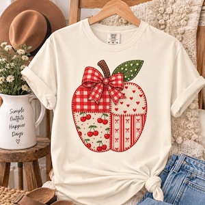 Puede incluir: Una camiseta de manga corta color crema con un gráfico grande de una manzana compuesta por cuatro secciones de patchwork distintas. El diseño de la manzana incluye cuadros vichy rojos y blancos, corazones pequeños, cerezas y rayas rojas.