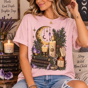 Może przedstawiać: Jasnoróżowy t-shirt z nadrukiem graficznym przedstawiającym półksiężyc, zapalone świece, fioletowe kryształy, gałązki rozmarynu i stos książek. Książki mają tytuły Moon Magic, Herbal Healing oraz A Brighter You, z dodatkowymi etykietami.