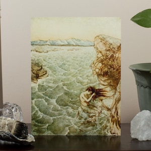 May include: An art print depicting a woman by the sea, with a rocky cliff on the right and mountains in the distance. The artwork features muted tones of green, brown, and beige, creating a serene and classical aesthetic.
