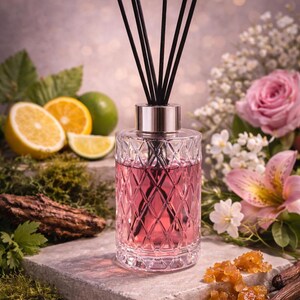 May include: A clear glass diffuser bottle with a pink liquid and black reeds. The bottle has a diamond pattern and a silver cap. The image includes citrus fruits, flowers, and decorative elements, suggesting a fragrance product.