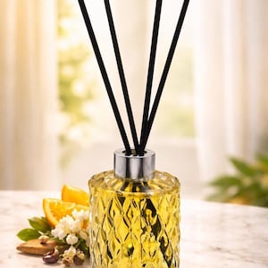 May include: A decorative glass diffuser bottle with a diamond pattern, filled with yellow liquid, and topped with a silver cap and black reeds. Sliced oranges and white flowers are arranged nearby, suggesting a citrus fragrance.
