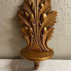 May include: A gold-colored wall shelf featuring an ornate, vertical back panel with carved acanthus leaf motifs. The shelf has a semi-circular flat surface for display and a small, decorative support bracket underneath the base of the shelf.
