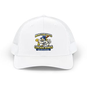 Op de afbeelding: Witte truckerpet met een mesh achterkant en een gebogen klep. De voorkant toont een cartoonkip in een honkbaluniform die met een knuppel zwaait, met de tekst "BEND YOUR KNEES & CHICKEN WINGS STAY LOW. SWING BIG."