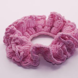 May include: A pink, handmade scrunchie with a textured, crocheted design. The scrunchie is a soft, light pink color and features a ruffled edge. This hair accessory is perfect for adding a touch of style to any hairstyle.
