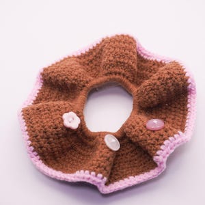 Könnte beinhalten: Ein braunes gehäkeltes Haargummi mit rosa Rand und drei dekorativen Knöpfen. Das handgefertigte Haaraccessoire hat eine Donut-Form. Die Knöpfe sind rosa und weiß und verleihen dem Haargummi Details. Durchmesser ca. 10 cm.