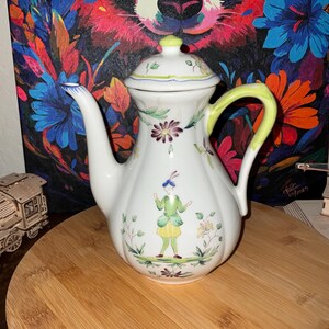 May include: White ceramic teapot with a lid, a curved spout, and a yellow handle. The teapot is decorated with floral and figure illustrations. The background features a colorful painting of a panda.