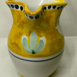 May include: A ceramic pitcher featuring a white base with a yellow painted upper section. The rim is decorated with blue dots, and the body displays a stylized light green floral motif. A thin blue horizontal line separates the yellow and white areas.