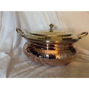 Puede incluir: Una olla redonda de cobre martillado con tapa de latón pulido y dos asas laterales de latón. La olla presenta un cuerpo ancho y redondeado con una superficie texturizada, mientras que la tapa incluye un pequeño pomo central puntiagudo.