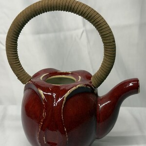 May include: A glossy, deep red ceramic teapot with a rounded body and a curved spout. The teapot features a wide, circular opening at the top and a tall, arched handle wrapped in a textured, tan-colored material for a secure grip.