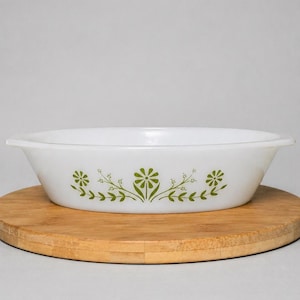 Puede incluir: Fuente ovalada blanca con diseño floral verde. La fuente tiene una forma sencilla con dos asas. El patrón floral incluye flores y hojas estilizadas. La fuente está sobre una tabla de cortar redonda de madera.