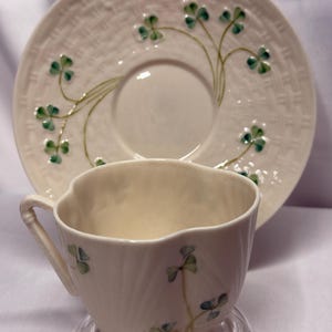 May include: A cream-colored ceramic teacup and matching saucer set. The saucer features a raised basket-weave texture and painted green shamrock motifs. The teacup has a scalloped rim, a curved handle, and matching green shamrock illustrations.